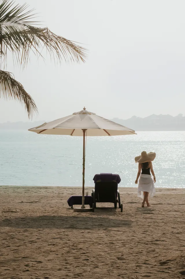 A woman in a wide-brimmed hat walks along the beach past a parasol and sun lounger.