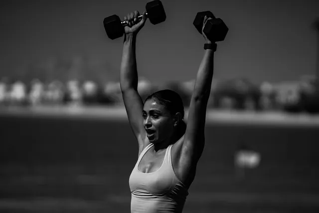 A woman presses dumbbells overhead, in black and white.