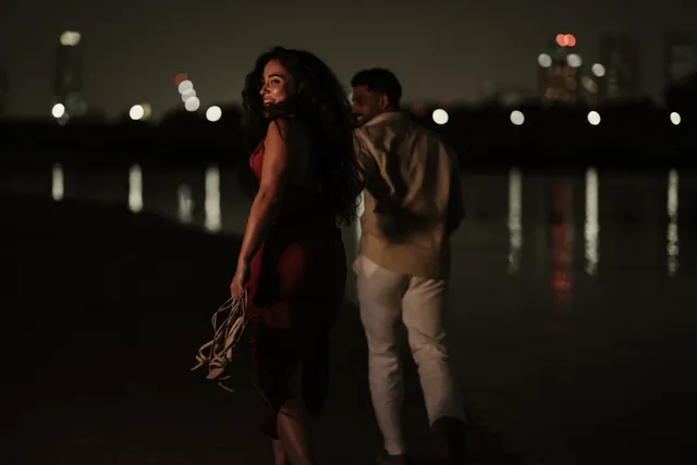 A couple walk along the shoreline at night with the city lights behind them.