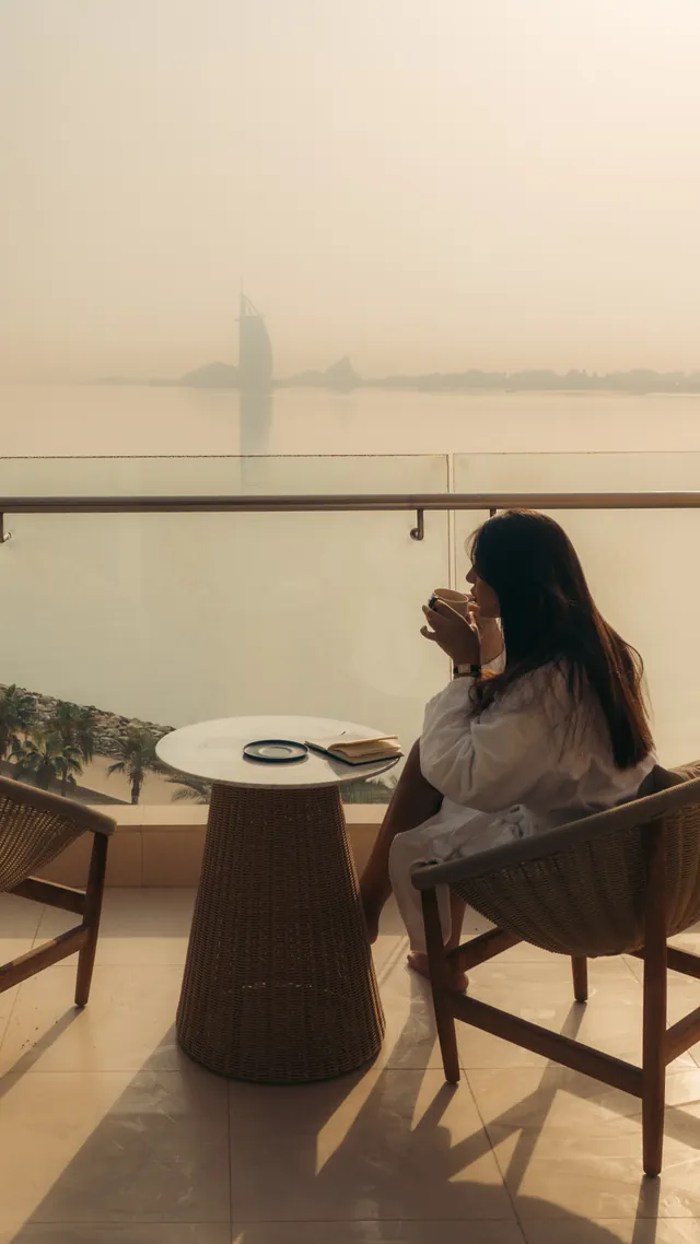 A woman drinks coffee on a terrace overlooking the sea and the Burj Al Arab.
